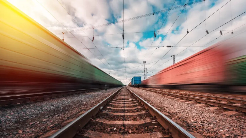 Ground-level view between railway tracks as two blurred freight trains speed past in opposite directions under a cloudy sky