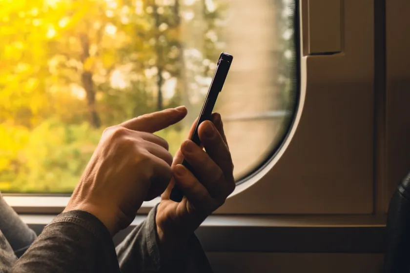 Hands holding a phone by a train window with autumn trees blurred outside