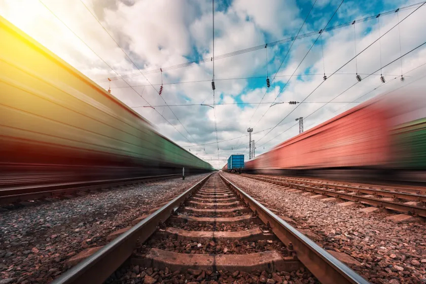 Ground-level view between railway tracks as two blurred freight trains speed past in opposite directions under a cloudy sky